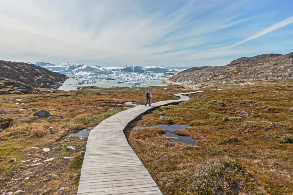 Tourist walking on a wooden pathway in the scenic Sermermiut Eskimo Settlement near Ilulissat, Greenland, with icebergs in the background.