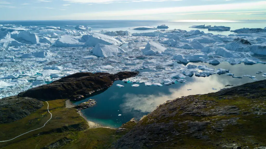 Aerial view of Ilulissat Icefjord in Greenland, showcasing numerous icebergs floating in glacial waters near rocky terrain, under a clear sky, ideal for Arctic travel and tourism.