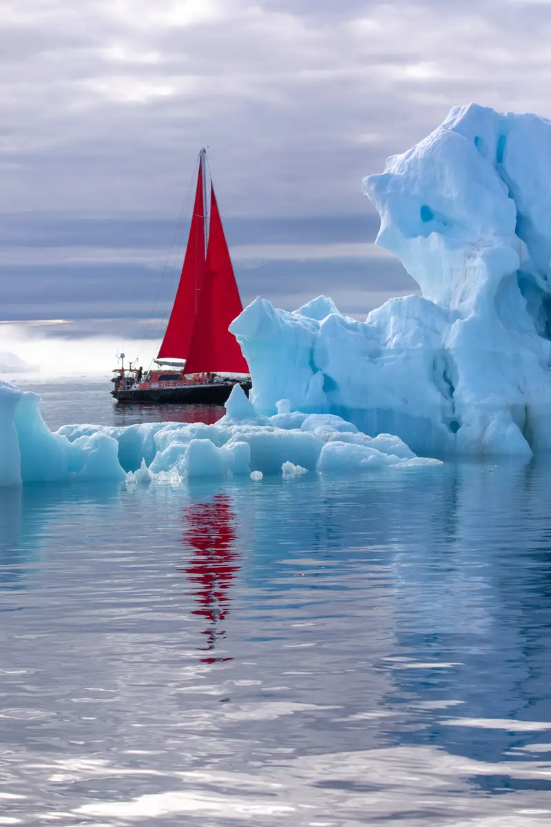 Red sailboat sailing beside a towering iceberg in Disko Bay, Ilulissat, Greenland, showcasing Arctic adventure tourism.