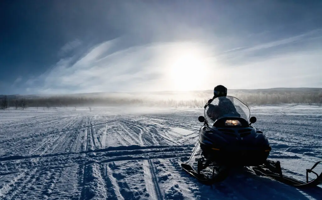 Person preparing to ride a snowmobile in snowy Lapland, Sweden, with sun halo effect over frosty landscape and birch forest in Hemavan-Tarnaby area.
