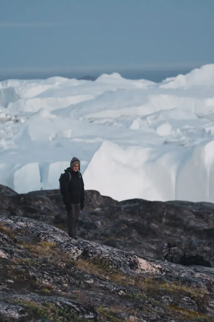 Tourist in warm clothing overlooking the vast Icefjord in Ilulissat, Greenland during twilight, showcasing the stunning arctic landscape and icebergs.