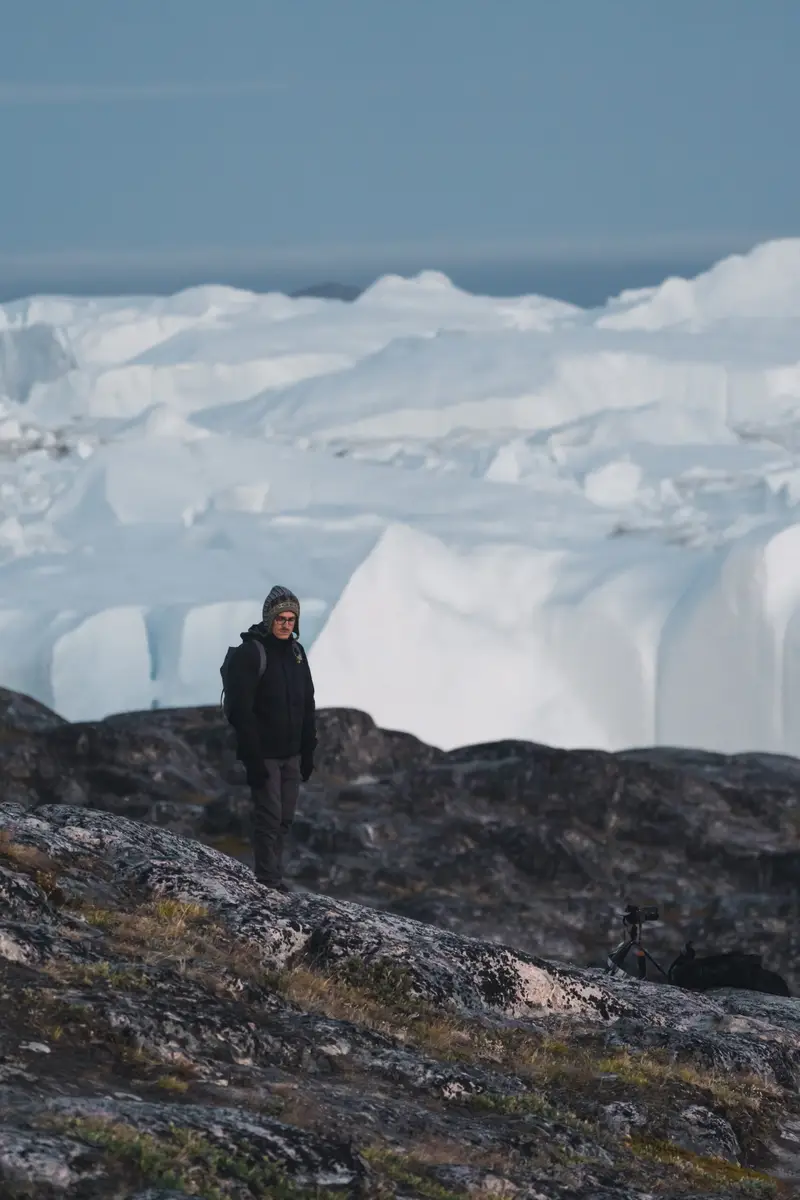 Tourist in warm clothing overlooking the vast Icefjord in Ilulissat, Greenland during twilight, showcasing the stunning arctic landscape and icebergs.