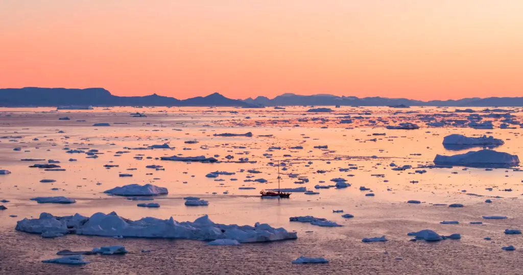 Breathtaking sunset over the Icefjord in Greenland, featuring a solitary boat amidst scattered icebergs in serene arctic waters.