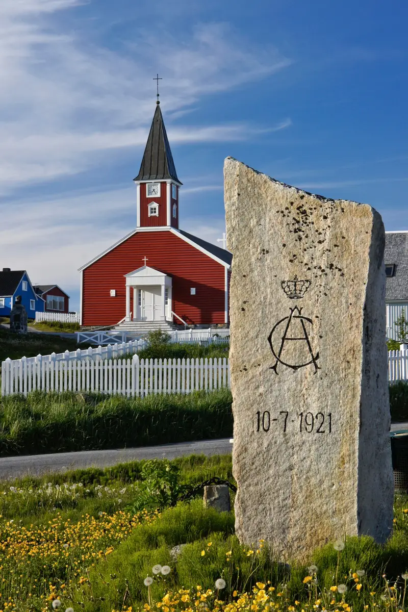 Historical commemorative stone with date and royal insignia in front of the vibrant red Frelserens Kirke in Nuuk, Greenland, marking the 1921 royal visit.